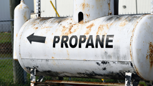 Photo of a large, rusted propane tank with the word "Propane" and an arrow painted on it for Applied Worldwide blog "Why Do Propane Tank Prices Vary So Much?"